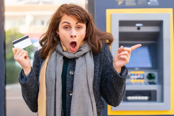 Brunette Woman Holding Credit Card Outdoors Surprised Pointing Finger Side — Stock Photo, Image