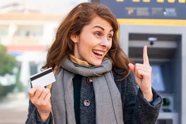 Brunette woman holding a credit card at outdoors intending to realizes the solution while lifting a finger up