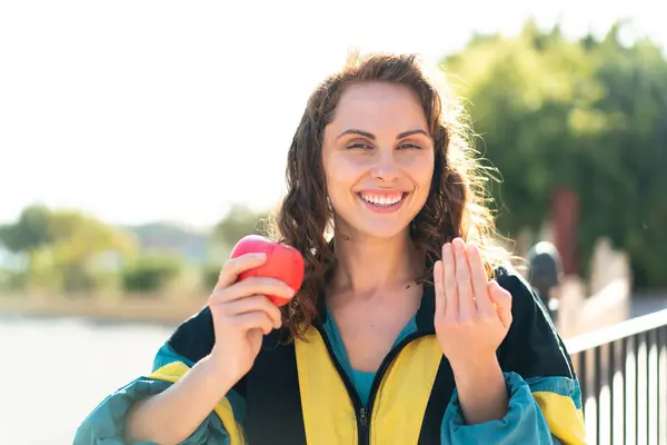 Young sport woman with an apple at outdoors inviting to come with hand. Happy that you came