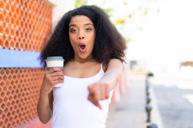 Genç Afrikalı Amerikalı bir kadın açık havada kahve tutuyor. Şaşırmış ve ön tarafı işaret ediyor.