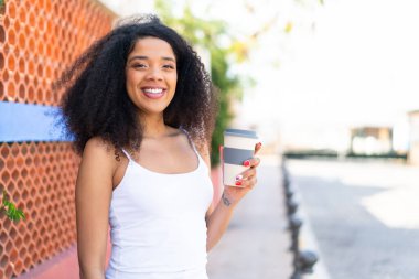 Genç Afrikalı Amerikalı bir kadın açık havada kahvesini yudumlarken gülümsüyor.