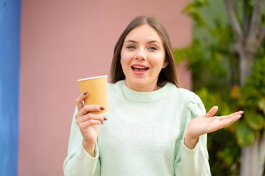 Sarışın, güzel bir kadın açık havada kahve tutuyor. Yüzünde şok ifadesi var.