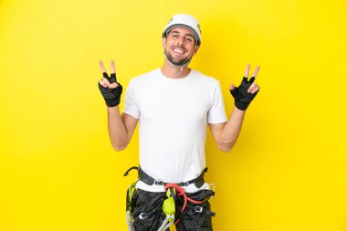 Sarı arka planda izole edilmiş kaya tırmanışçısı iki eliyle zafer işareti gösteriyor.