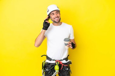 Genç kaya tırmanışçısı sarı arka planda izole edilmiş. Elinde kahve ve bir cep telefonu var.