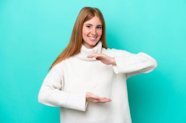 Mavi arka planda izole edilmiş genç kızıl saçlı kadın bir ilan vermek için avucunun içinde hayali bir şey tutuyor.