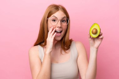 Elinde avakado tutan genç kızıl saçlı kadın. Pembe arka planda, şaşırmış ve şok olmuş bir yüz ifadesi var.