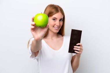 Genç kızıl saçlı kadın beyaz arka planda izole edilmiş. Bir elinde çikolata tableti, diğer elinde elma var.