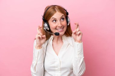 Tele pazarlamacı kızıl saçlı kadın pembe arka planda izole edilmiş kulaklıkla çalışıyor.