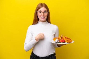 Sarı arka planda, yüzünde sürpriz bir ifade olan waffle tutan genç kızıl saçlı kadın.