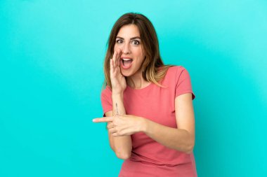Mavi arka planda izole edilmiş genç beyaz kadın bir ürünü sunmak ve bir şeyler fısıldamak için kenarı işaret ediyor.