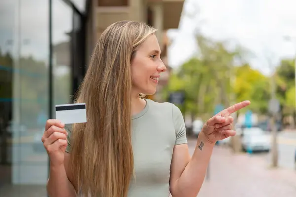 Young blonde woman holding a credit card at outdoors pointing to the side to present a product