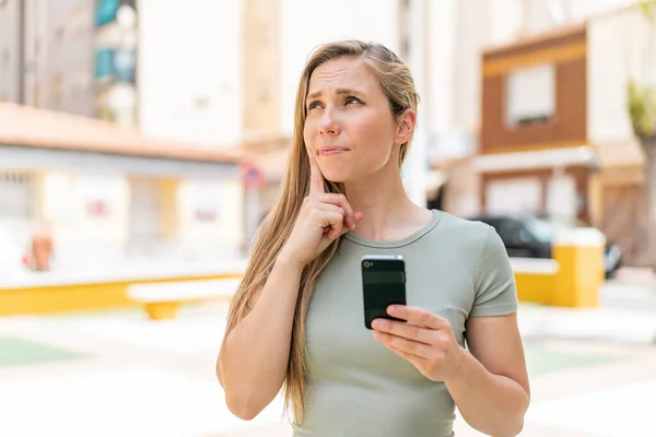 Young Blonde Woman Outdoors Using Mobile Phone Thinking — Stock Photo, Image
