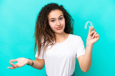 Mavi arka planda izole edilmiş görünmez diş telleri tutan genç bir Arap kadını omuzlarını kaldırırken şüpheli hareketlerde bulunuyor.