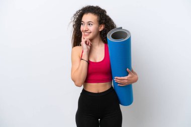 Genç Arap spor kadını yoga derslerine giderken beyaz arka planda izole edilmiş bir minder tutuyor ve yukarı bakarken bir fikir düşünüyor.
