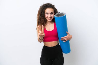 Genç Arap spor kadını yoga derslerine giderken beyaz arka planda izole edilmiş bir minder tutuyor gülümsüyor ve zafer işareti gösteriyor.