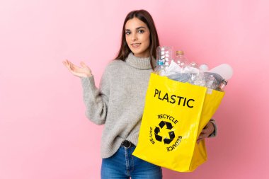 Pembe arka planda izole edilmiş plastik şişelerle dolu bir çanta taşıyan beyaz bir kadın davet edilmek için ellerini yana uzatıyor.