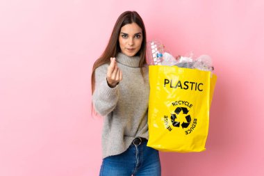Pembe arka planda izole edilmiş plastik şişelerle dolu bir çanta taşıyan beyaz bir kadın para hareketi yapıyor.