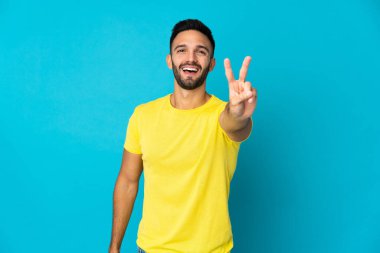 Mavi arka planda izole edilmiş genç beyaz adam gülümsüyor ve zafer işareti gösteriyor.