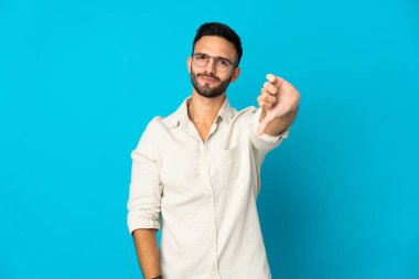 Mavi arka planda izole edilmiş genç beyaz adam olumsuz ifadeyle baş parmağını aşağı gösteriyor.