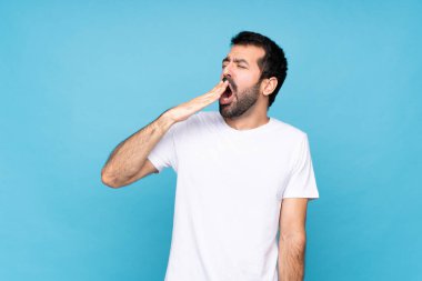 Sakallı genç adam izole edilmiş mavi arka planda esniyor ve ağzını eliyle açık tutuyor.