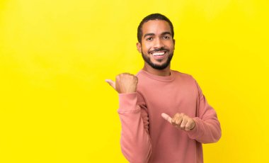 Sarı arka planda izole edilmiş genç latin adam bir ürün sunmak için kenarı işaret ediyor.