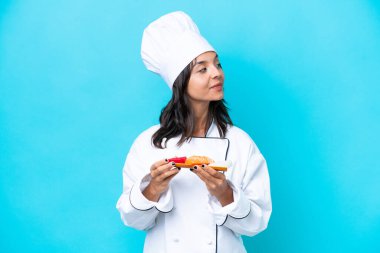 Mavi arka planda saşimi tutan genç İspanyol aşçı kadın yan tarafa bakıyor.