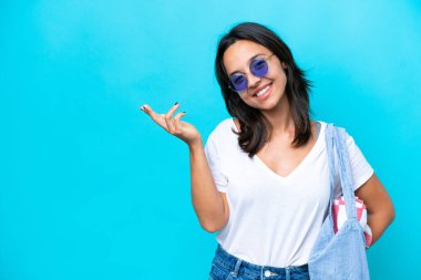 Elinde kumsal çantası olan genç beyaz bir kadın mavi arka planda izole edilmiş ve davet edilmek için ellerini yana uzatıyor.