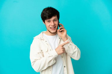 Mavi arka planda izole edilmiş cep telefonu kullanan genç Rus bir ürünü sunmak için kenarı işaret ediyor.