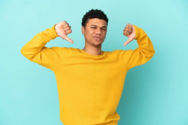 Mavi arka planda izole edilmiş genç Afrikalı Amerikalı iki eliyle baş parmağını gösteriyor.