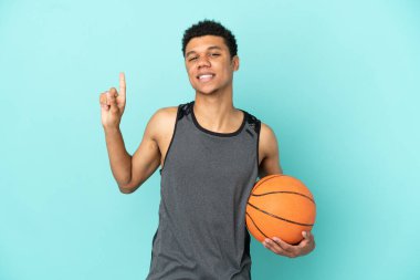 Basketbolcu Afro-Amerikan adam mavi arka planda izole edilmiş ve en iyinin işareti olarak parmağını kaldırıyor.