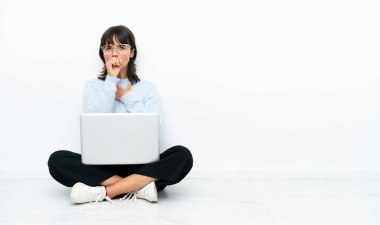 Beyaz arka planda dizüstü bilgisayarla yerde oturan genç melez kadın öksürüp kendini kötü hissediyor.