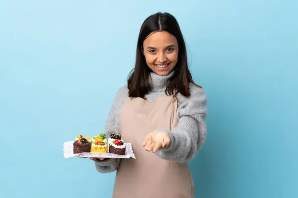 Pastry chef holding a big cake over isolated blue background holding copyspace imaginary on the palm to insert an ad.
