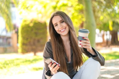 Dışarıda cep telefonu kullanan ve elinde kahve tutan genç bir kadın.