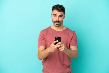 Mavi arka planda izole edilmiş genç beyaz adam şaşırdı ve bir mesaj gönderiyor.