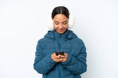 Kışlık kulaklık takan genç İspanyol kadın beyaz arka planda izole edilmiş cep telefonuyla bir mesaj gönderiyor.