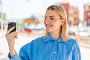 Açık havada cep telefonu kullanan güzel, sarışın bir kadın. Mutlu bir ifadeyle.