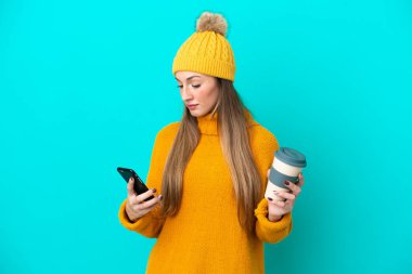 Kışlık ceket giyen beyaz bir kadın mavi arka planda izole edilmiş. Elinde kahve ve cep telefonu var.
