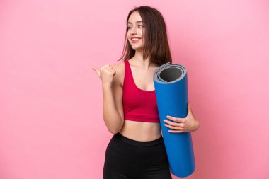 Genç sporcu Ukraynalı kadın yoga derslerine gidiyor. Elinde pembe arka planda izole edilmiş bir minder var. Ürünü sunmak için yan tarafa bakıyor.