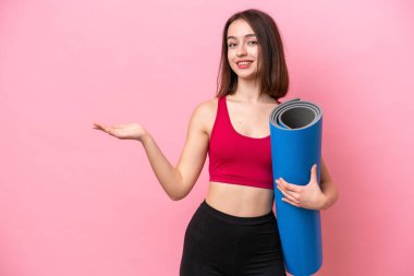 Young sport Ukrainian woman going to yoga classes while holding a mat isolated on pink background extending hands to the side for inviting to come