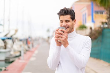 Açık havada genç beyaz bir adam elinde neşeli bir ifadeyle kahveyle geziyor.