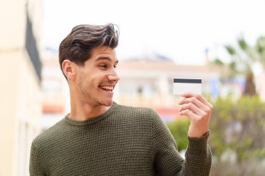 Açık havada elinde mutlu bir ifadeyle kredi kartı tutan genç beyaz bir adam.