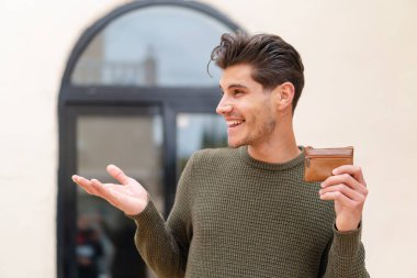 Açık havada cüzdan tutan genç beyaz bir adam. Yüzünde sürpriz bir ifade var.