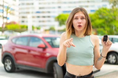Dışarıda arabanın anahtarlarını tutan genç sarışın kadın şaşırmış ve ön tarafı işaret ediyor.