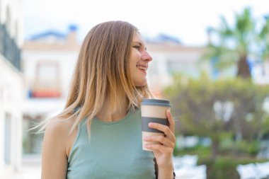 Sarışın bir kadın açık havada elinde kahveyle neşeli bir ifade ile bekliyor.
