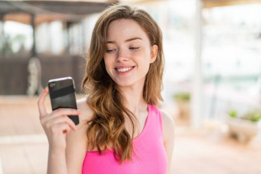 Genç kızıl saçlı kadın açık havada mutlu bir ifadeyle cep telefonu kullanıyor.