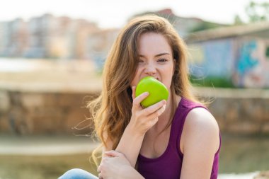 Dışarıdaki kızıl saçlı genç kadın elma yiyor.