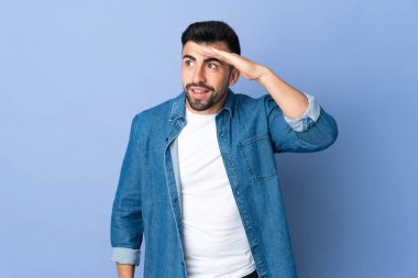 İzole edilmiş mavi arka planı olan beyaz bir adam, bir şeye bakmak için elini uzaklara bakıyor.