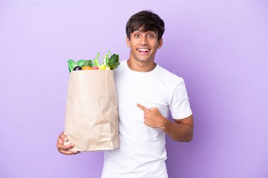 Elinde bir alışveriş çantası tutan genç adam mor arka planda, şaşırmış bir yüz ifadesiyle izole edilmiş.