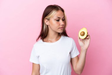 Avokado tutan genç Rus kadın pembe arka planda izole edilmiş, yan tarafa bakıyor.