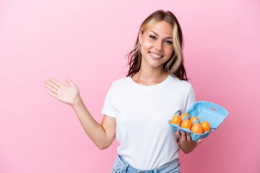 Pembe arka planda yumurtaları tutan genç Rus kadın davet edilmek için ellerini yana uzatıyor.